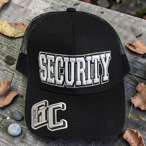 Op de afbeelding: Zwarte truckerpet met het woord "SECURITY" geborduurd in witte blokletters. De pet heeft ook de letters "GC" in een zilver en wit ontwerp op de voorkant. De pet heeft een zwarte mesh achterkant.