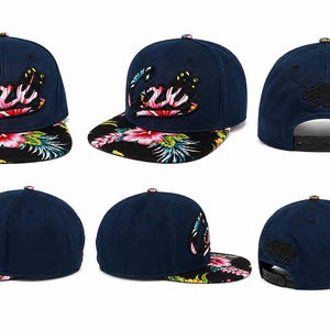 May include: A navy blue snapback cap featuring the word Cali embroidered in a floral pattern on the front. The flat brim displays a black background with a vibrant pink, green, and white tropical hibiscus flower and leaf print. A black bear silhouette is embroidered on the back.