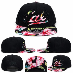 May include: A black snapback cap featuring the word Cali embroidered in black on the front crown. The flat brim displays a vibrant floral pattern with pink hibiscus flowers and green leaves. The back panel includes a small black bear silhouette.
