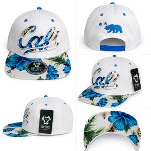 May include: A white snapback cap featuring the word Cali in white script with a blue floral pattern fill. The flat brim displays a vibrant blue hibiscus and green palm leaf print. The back includes a blue bear and star embroidery with a snap closure.