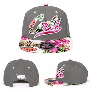May include: A gray snapback hat featuring the word Cali in white script embroidery filled with a pink and green floral pattern. The flat brim displays a vibrant tropical hibiscus print. The back shows a white bear silhouette and a star.