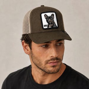 May include: An olive green baseball cap with a mesh back and a square patch on the front. The patch features a black and white illustration of a French Bulldog with the word DOG printed in black capital letters below the dog's chin.