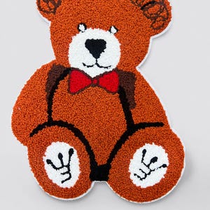 Op de afbeelding: Een getuft tapijt in de vorm van een teddybeer met roestoranje vacht, een witte snuit en een zwarte neus. De beer heeft een felrode vlinderdas, zwarte gestikte contouren en witte ronde vlekken op de voeten met zwarte pootafdrukken.