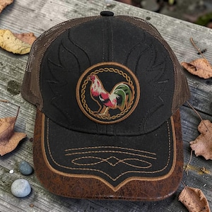 Pode incluir: Boné de caminhoneiro preto com parte traseira em malha castanha e aba castanha com efeito de couro. O boné apresenta um patch circular com um desenho de galo em vermelho, verde e dourado. O boné tem costuras decorativas e uma aba curva.