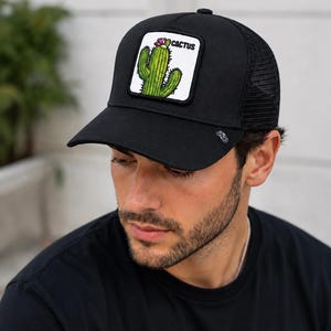 Puede incluir: Una gorra tipo camionero de color negro con un parche cuadrado blanco en el panel frontal. El parche muestra una ilustración de un cactus verde con una pequeña flor rosa y la palabra CACTUS impresa en negro. La gorra tiene visera negra y malla.