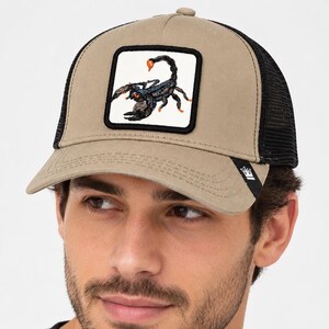 Peut inclure: Une casquette de baseball beige avec un arrière en filet noir et un écusson carré sur le devant. L'écusson présente une illustration détaillée d'un scorpion noir avec des accents orange sur fond blanc. Une petite étiquette noire avec un logo couronne blanc est sur la visière.