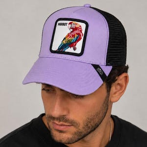 Puede incluir: Una gorra tipo trucker de color lavanda con malla negra en la parte posterior y un parche cuadrado en el frente. El parche muestra una ilustración colorida de un loro con la palabra PARROT impresa en negro sobre el ave. La visera es lavanda.