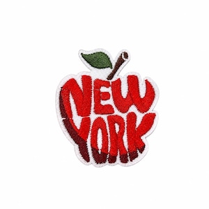 May include: An embroidered patch shaped like an apple with the text NEW YORK in bold red letters. The apple features a green leaf and a brown stem, with a white border outlining the entire shape against a plain white background.