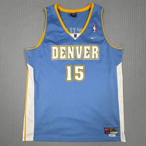 May include: A light blue basketball jersey featuring the word DENVER and the number 15 in white with yellow outlines. The sleeveless shirt has yellow trim around the neck and armholes, white side panels, and a small white Nike swoosh logo.