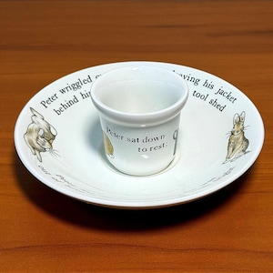 May include: A white ceramic egg cup and saucer set featuring illustrations of a rabbit and black text. The saucer reads Peter wriggled out, leaving his jacket behind him in the tool shed. The egg cup reads Peter sat down to rest.
