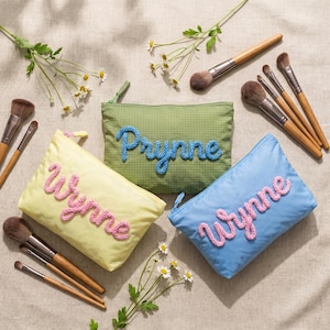 May include: Three cosmetic bags in yellow, green, and blue, each with a name embroidered in a contrasting color. Makeup brushes and sprigs of white flowers are arranged around the bags, suggesting a beauty theme.