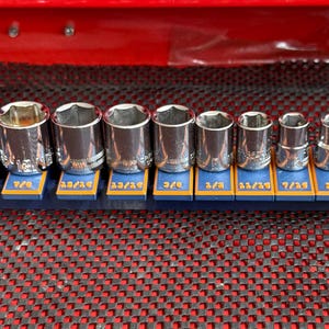May include: A set of twelve silver-colored metal socket tools arranged in descending order of size on a blue plastic rail. Each socket is positioned above a blue and orange label displaying its specific imperial measurement in inches.
