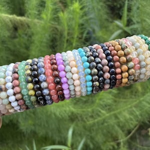 May include: A collection of beaded bracelets in various colors, including orange, green, black, and purple. The bracelets are displayed on a cylindrical, beige surface against a backdrop of green foliage. Each bracelet is made of small, round beads.