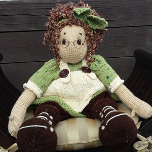 Puede incluir: Una muñeca de punto con cabello castaño, vestida con ropa verde y blanca, sentada en una silla de madera marrón. La muñeca lleva gafas y botas marrones.