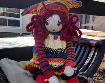 May include: A colorful knitted doll with red hair, wearing a striped sweater, a patterned skirt, and a multi-colored hat. The doll is sitting on a wooden bench with a striped cushion.