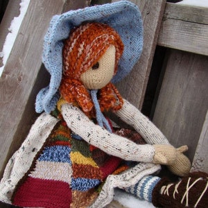 May include: A knitted doll wearing a blue and white hat, a patchwork dress, and brown boots. The doll is sitting on a wooden surface with snow.