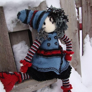 May include: A knitted doll with a blue and black striped hat, a blue and red striped dress, and red boots. The doll is sitting on a wooden fence post in the snow.