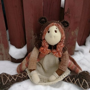 Può includere: Una bambola fatta a mano all'uncinetto con cappuccio e orecchie da orso, seduta sulla neve. La bambola presenta un viso e un corpo color crema, con dettagli marroni e arancioni sul cappuccio, sulle braccia e sulle gambe. Le gambe della bambola sono distese, contro una staccionata di legno rossa.