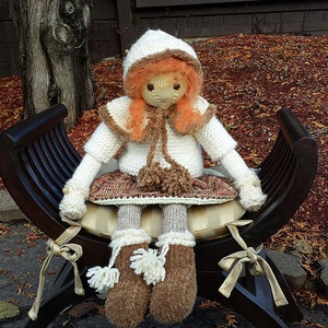 May include: A knitted doll wearing a white hooded sweater, brown boots, and a brown skirt sits on a wooden bench with a white cushion. The doll has red hair and brown eyes.