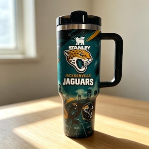 May include: A teal and black Stanley tumbler with a handle and lid, featuring the Jacksonville Jaguars logo, the text "STANLEY" and "JACKSONVILLE JAGUARS," and a graphic of a football field and players printed on the exterior surface.