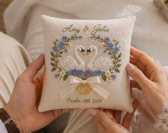 May include: A square cream-colored ring bearer pillow featuring embroidered white swans facing each other, surrounded by a floral wreath of blue and green flowers. A white ribbon bow holds two gold rings, with the text Amy & John and October 19 2026.