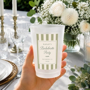 May include: A frosted plastic cup featuring a sage green and white vertical striped label with the text Kailey's Bachelorette Party, San Diego, CA, 2026. The cup has a smooth, translucent finish and is held upright against a blurred floral background.