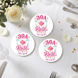 Puede incluir: Tres posavasos redondos blancos con un gráfico rosa de una copa de vino llena con un atardecer y olas, flanqueada por palmeras. El texto dice 30A ROSÉ STATE OF MIND en letras rosas y verde azulado, con un pequeño corazón como detalle.