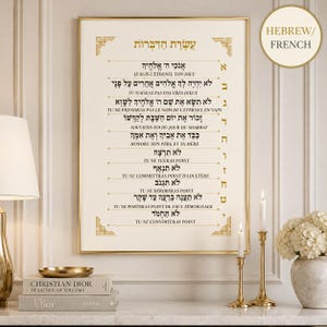 Könnte beinhalten: Ein gerahmter Druck mit hebräischen und französischen Texten. Der Druck hat einen goldenen Rahmen und zeigt die Zehn Gebote. Der Text ist in Schwarz und Gold gehalten. Der Druck wird auf einer weißen Oberfläche mit einer Lampe, Büchern und Kerzen angezeigt.