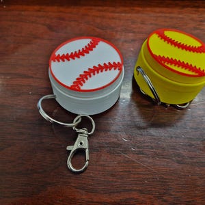 Puede incluir: Dos llaveros con temática de béisbol. Uno es blanco con costuras rojas, el otro es amarillo con costuras rojas. Ambos tienen forma redonda y un clip plateado para sujetarlos a llaves o bolsos. Los llaveros están sobre una superficie de madera oscura.