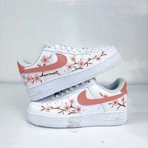 以下が含まれることがあります： ピンクの花と茶色の枝が手描きされた桜のデザインが施された、白いローカットスニーカー。サイドにはダスティローズカラーのスウッシュロゴ、白い靴紐、そしてAirの刻印が入った厚みのある白いラバーソールが特徴です。