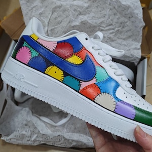 May include: White Air Force 1 sneakers with a colorful patchwork design on the side panels. The design features various colors including blue, red, yellow, pink, and green. The Nike swoosh is blue.