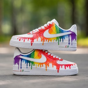 Puede incluir: Un par de zapatillas blancas de corte bajo con un vibrante patrón de goteo de arcoíris en los laterales. Los zapatos tienen cordones blancos, un logotipo de Nike blanco y el texto AIR impreso en letras multicolores en las suelas blancas.