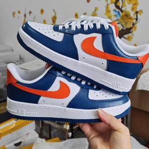 May include: A pair of white, blue, and orange sneakers. The shoes have a white sole, white laces, and a blue and white upper with an orange swoosh. The word "AIR" is visible on the side of the white sole.