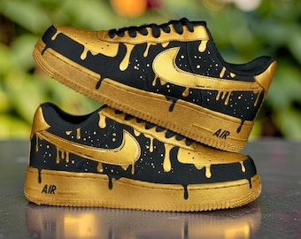 Op de afbeelding: Een paar zwarte en gouden op maat gemaakte sneakers. De schoenen hebben een zwarte basis met gouden accenten, waaronder een druipend verfdesign en de Nike swoosh. Het woord "AIR" is zichtbaar op de zijkant van de schoen.