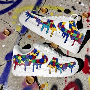 May include: White sneakers with colorful puzzle piece designs and dripping paint accents. The shoes feature a classic silhouette with a low profile, white laces, and a rubber sole. The design incorporates red, yellow, blue, green, and purple puzzle pieces.