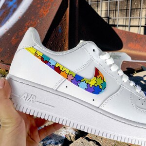 May include: White leather sneaker with a colorful puzzle piece design on the side and a matching puzzle piece Nike swoosh. The shoe has white laces and a white sole. The word "AIR" is visible on the side of the sole.
