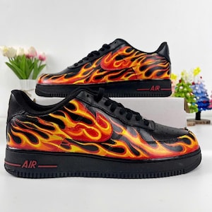 May include: Pair of black sneakers with a vibrant flame design in shades of red, orange, and yellow. The sneakers have a black sole with the word "AIR" in red. The design covers the sides and toe of the shoes.