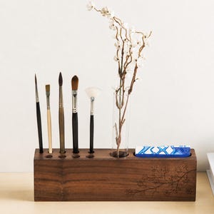 May include: A dark brown wooden desk organizer with holes for paint brushes and a glass vase holding white flowers. A small blue and white box sits in a slot. The organizer has an etched floral design.