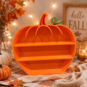 May include: A bright orange pumpkin-shaped shelf with two horizontal tiers for display. The shelf features a curved top edge and a small, distinct green curly stem attached to the upper right side, creating a functional storage piece.