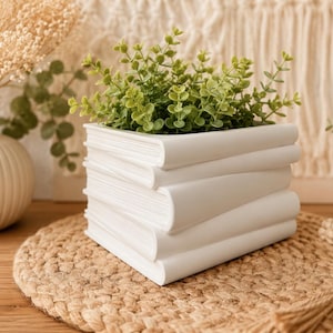 May include: A white ceramic planter shaped like a stack of three books. The planter has a square opening at the top and contains a small green artificial plant with rounded leaves. The exterior surface is smooth and matte, mimicking book spines.