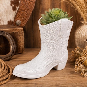 May include: A white ceramic cowboy boot planter featuring an intricate embossed scrollwork pattern across the entire surface. The boot has a classic silhouette with a low heel and a pull tab at the top, currently holding a green succulent plant.