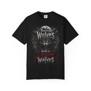 Può includere: T-shirt nera con un disegno di lupo e il testo "Raised by Wolves" e "Asked to Leave by Wolves". Il design presenta un lupo, artigli e una luna piena in tonalità grigie e rosse.