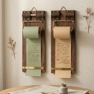 May include: Two wall-mounted paper roll dispensers featuring dark wood rectangular backings, brass hardware, and brown leather straps. Each unit holds a roll of paper with handwritten cursive text, one in sage green and one in light tan.