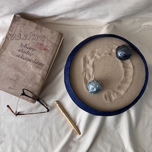Op de afbeelding: Een vintage boek getiteld VARÍME zdravo chutno a hospodárne, een bril met bruin montuur, een goudkleurige pen en een ronde blauwe bak gevuld met zand met twee metalen bollen en geharkte patronen in het zandoppervlak.