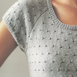 Può includere: Un top in maglia grigio chiaro con scollatura rotonda e maniche corte. Il tessuto è adornato da piccole perline grigio scuro, spaziate uniformemente. La trama e le perline del capo creano un design sottile ed elegante.