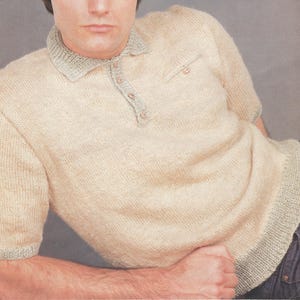 May include: A short-sleeved knit polo shirt in a light beige color with a contrasting grey collar, sleeve cuffs, and bottom hem. The shirt features a button-up placket with four small buttons and a single small pocket on the upper left chest area.