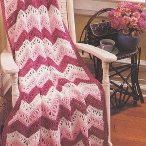 May include: A crochet blanket featuring a chevron pattern in alternating stripes of dark magenta, light pink, and cream. The textile has an openwork stitch design with a scalloped edge, draped over a white wooden chair with vertical slats.