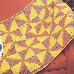Peut inclure: Une couverture crochetée à la main présentant un motif récurrent de moulinet. Le design se compose de sections triangulaires jaune vif alternant avec des blocs texturés de fil panaché dans des tons orange, violet et marron, avec une bordure.
