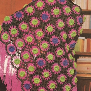May include: A crochet shawl featuring a repeating pattern of flower motifs in bright pink, lime green, and dark purple, connected by black yarn. The triangular garment is finished with long, vibrant pink fringe along the bottom edges.