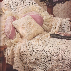 May include: An off-white crocheted throw blanket with intricate floral patterns draped over a surface, accompanied by two pink pillows with lace trim, a square crocheted pillow, a dark book with gold lettering, and a vase of purple flowers.